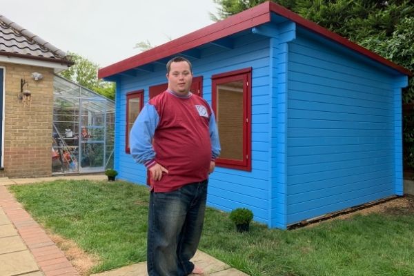 Man stands beside small blue shed with red trim on grassy lawn in backyard