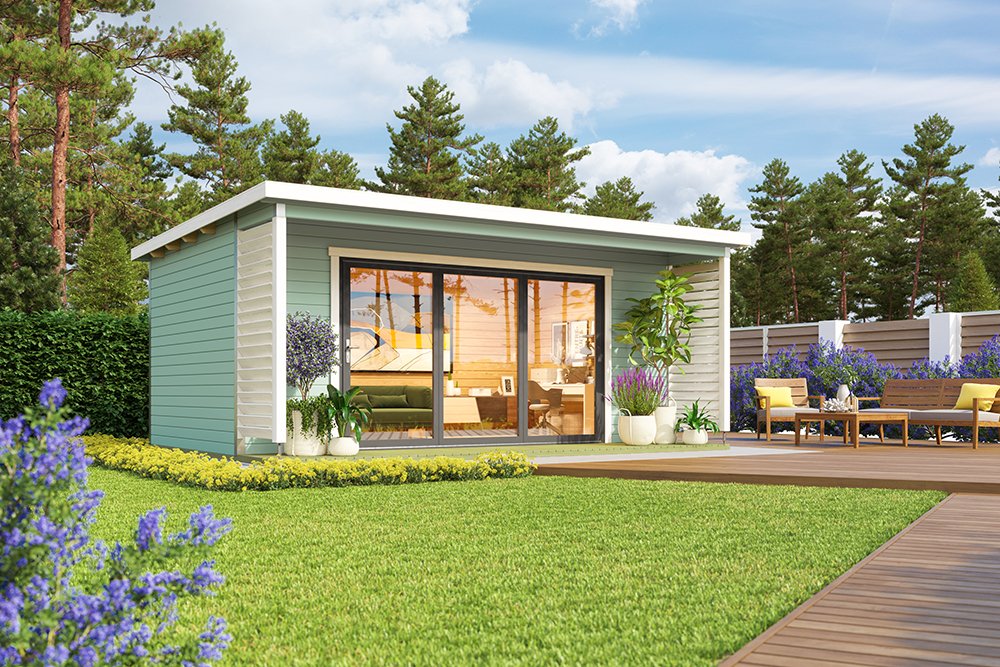 A small green shed with white trim and large glass doors sits on a deck surrounded by grass, flowers, and trees on a sunny day.