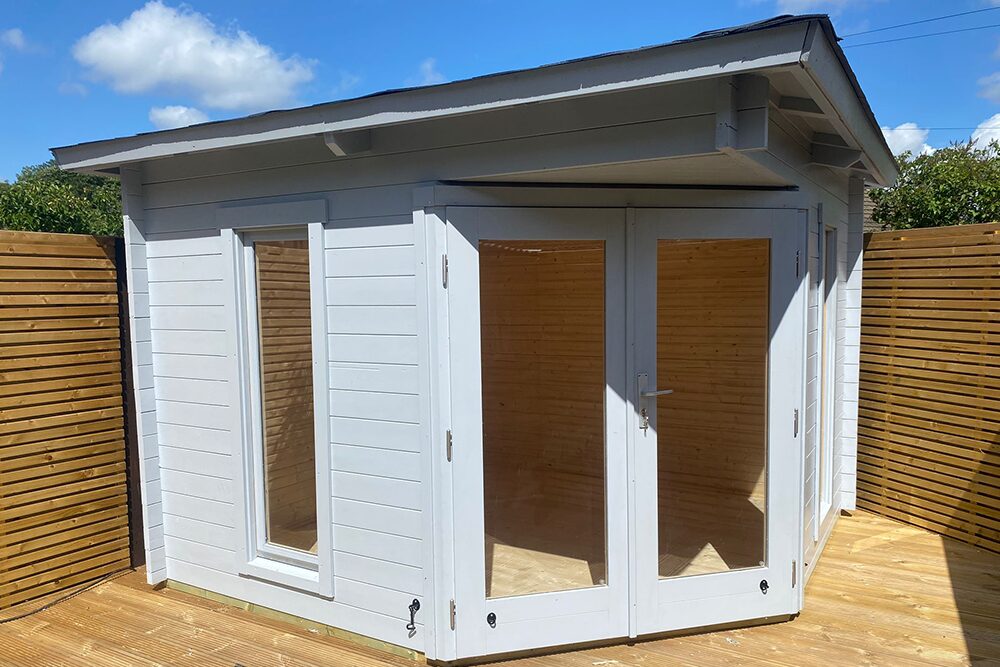 A white wooden shed with glass doors and a sloping roof on a deck surrounded by a brown wooden fence and trees under a blue sky