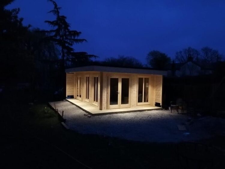 A small wooden cabin with large windows and doors lit up at night on a gravel base surrounded by darkness and trees