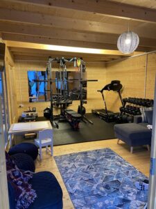 A log cabin home gym with wood walls and ceiling, exercise equipment, and a treadmill on black mats, with a blue rug and furniture.