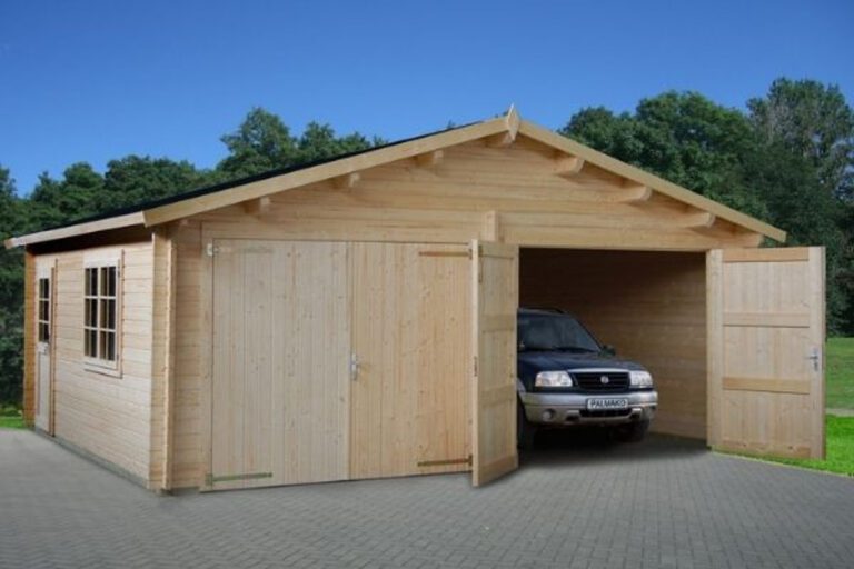 A light wooden garage with a car inside on a grey brick driveway, surrounded by trees and a clear blue sky