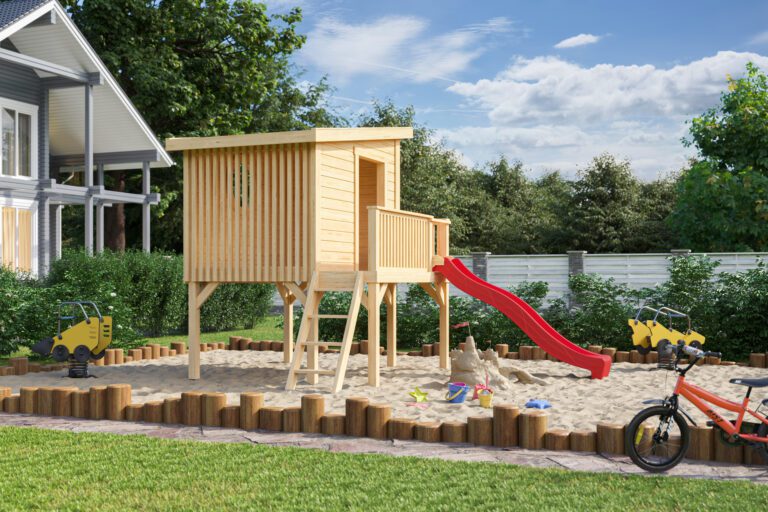 A wooden playhouse with a red slide in a sandbox surrounded by logs, with a house and trees in the background on a sunny day.
