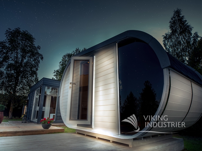 Modern white and black sauna cabin with curved roof and large window at night in a serene outdoor setting with trees