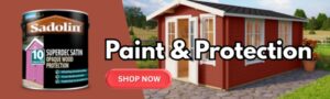 Red shed with white trim and black roof, surrounded by grass and trees, with a can of Sadolin paint