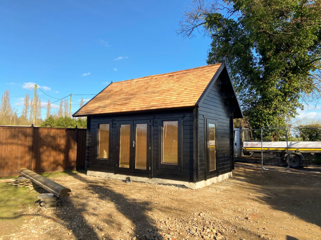 A small black shed with a brown roof and glass doors sits on a dirt lot, surrounded by a wooden fence and trees under a blue sky.