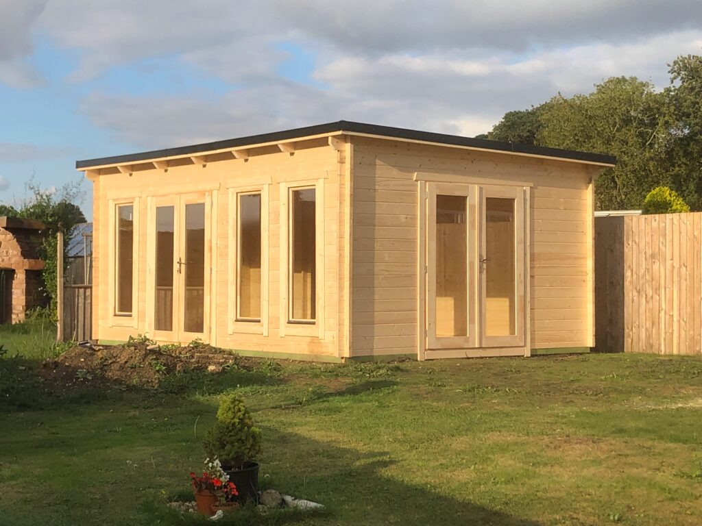 A light wooden shed with large windows and a door on a grassy lawn with a small plant and flowers, under a cloudy sky