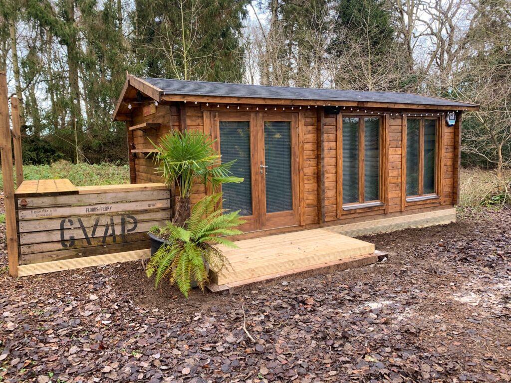 A small log cabin with brown wood and black roof in a wooded area with a wooden deck and plants