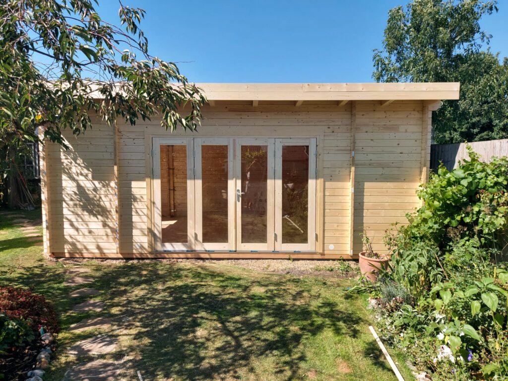 A light wooden shed with glass doors stands in a garden on a sunny day surrounded by trees and plants