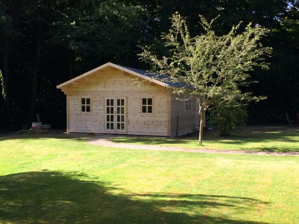 A small log cabin with light wood and white trim sits on green grass with a tree beside it.