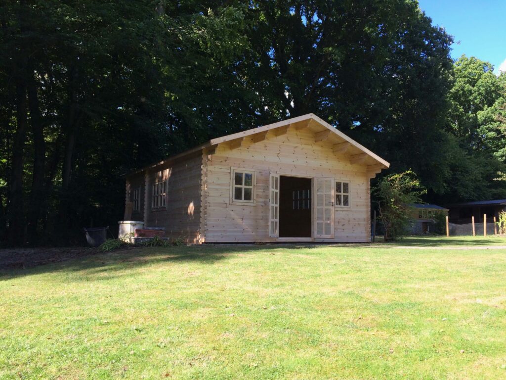 A small log cabin with light wood and a gable roof sits on grass with trees and a car port in the background on a sunny day