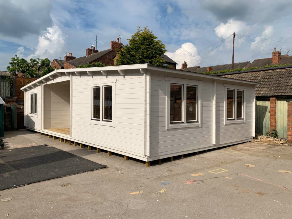 A white shed with a car port area and several windows sits on a concrete lot surrounded by houses and a cloudy blue sky.