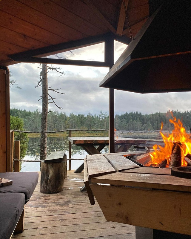 Cozy log cabin deck with fire pit, wooden furniture, and serene lake view under cloudy sky