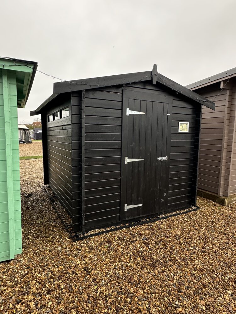 A small black shed with double doors and a peaked roof on gravel beside green and brown sheds